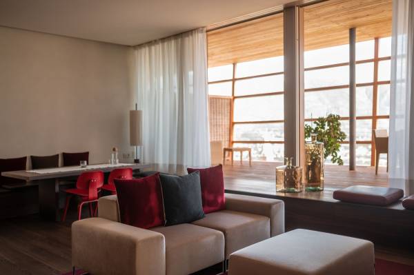 Moderne Hotelzimmer mit einem beigen Sofa, roten Kissen und roten Stühlen. Große Fenster mit Blick auf eine Terrasse und Pflanzen.