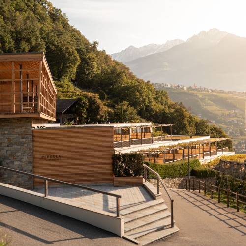 Moderne Hotelanlage mit Holz- und Steinfassade, umgeben von Bäumen und Weinreben. Im Hintergrund Berge und Sonnenuntergang.