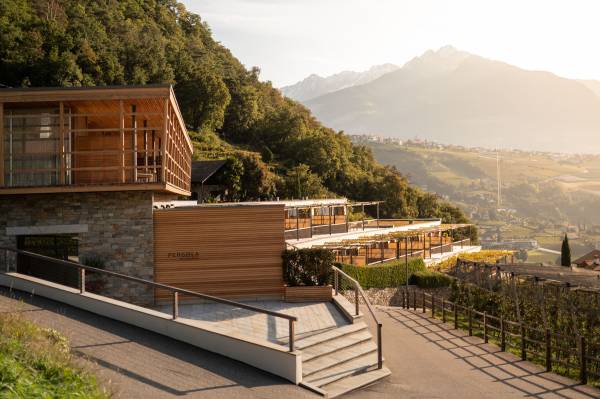 Moderne Hotelanlage mit Holz- und Steinfassade, umgeben von Bäumen und Weinreben. Im Hintergrund Berge und Sonnenuntergang.