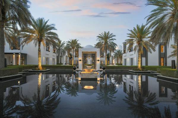 Panoramaansicht der Hotelanlage The Chedi Muscat mit Palmen, einem Wassergarten und modernen Gebäuden. Reflexionen im Wasser sind sichtbar.