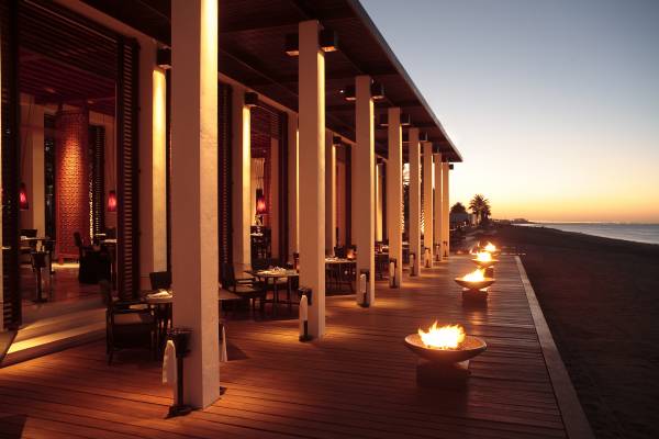 Die Hotelanlage 'The Chedi Muscat' zeigt ein Strandrestaurant mit Holzdeck, beleuchtet von Säulen und Feuerschalen, bei Sonnenuntergang.