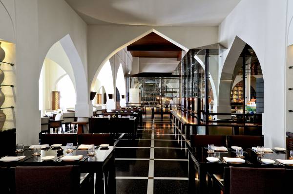 Innenansicht des Restaurants 'The Chedi Muscat' mit eleganten Tischen und Stühlen, hohen Decken und großen Bögen. Sichtbare Gläser und Geschirr auf den Tischen.