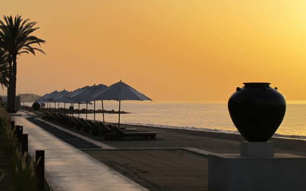 Strandansicht des Chedi Muscat bei Sonnenuntergang, mit Sonnenschirmen und Liegen, Palmen im Hintergrund, schwarzem Topf auf einem Podest.