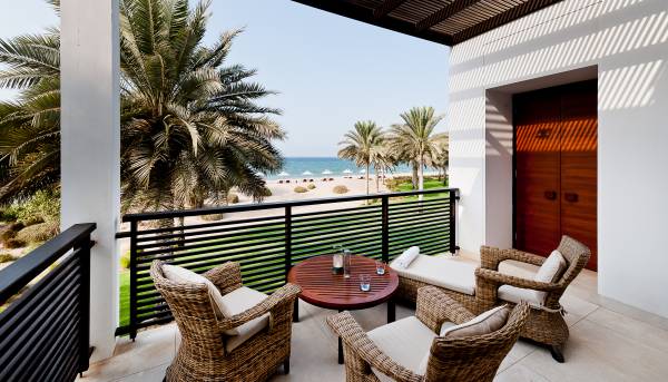 Balkon der Club Suite im Hotel The Chedi Muscat mit Meerblick. Zwei geflochtene Stühle und ein runder Tisch stehen auf dem Balkon. Im Hintergrund sind Palmen und der Strand sichtbar.