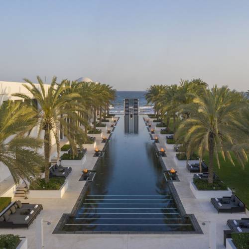 Blick auf den langen Pool der Hotelanlage The Chedi Muscat, umgeben von Palmen und Liegen. Im Hintergrund ist das Meer sichtbar.