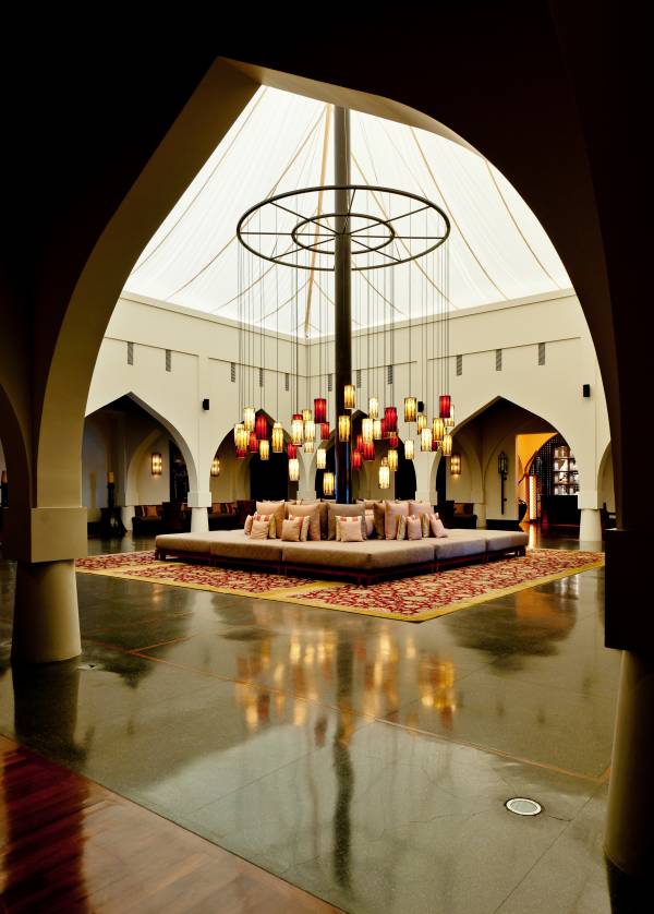 Lobby des Hotels 'The Chedi Muscat' mit einem großen, runden Sofa in der Mitte, umgeben von bunten Pendelleuchten und einem reflektierenden Boden.