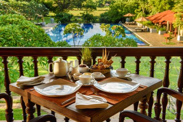 Fr&uuml;hst&uuml;ckstisch mit Tassen, Teekanne und Geb&auml;ck auf der Terrasse des Liyawela Hotels mit Blick auf den Pool und Garten