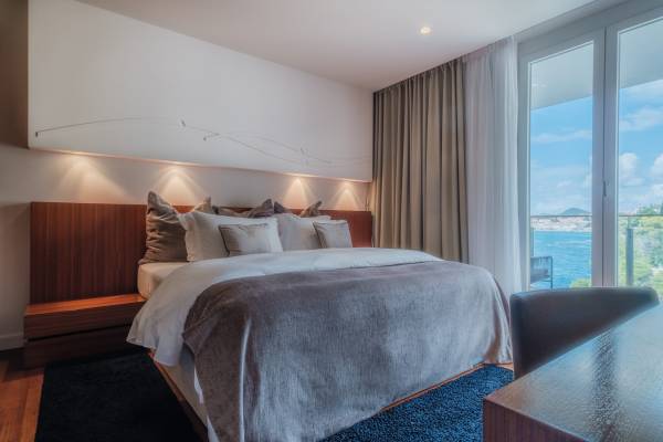 Standard-Suite mit großem Doppelbett, grauer Bettdecke, mehreren Kissen, Holzkopfteil und Blick auf Wasser durch bodentiefe Fenster