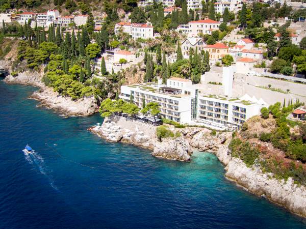 Hotelgebäude an felsiger Küste mit Blick auf das blaue Meer, umgeben von Bäumen und weiteren Häusern auf einem Hügel