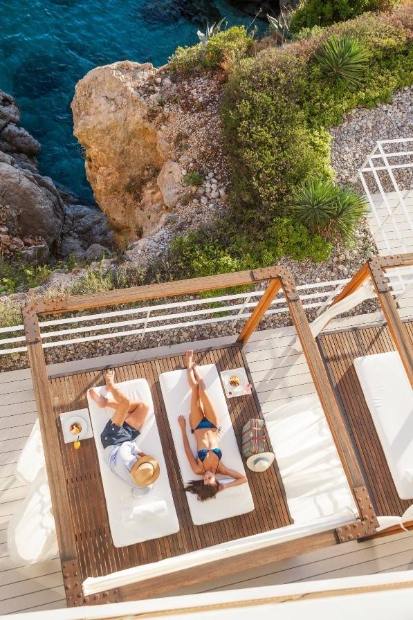 Zwei Personen entspannen auf Liegen unter einer Holzpergola mit Blick auf Felsen und Meer, Hotelumgebung mit mediterraner Vegetation