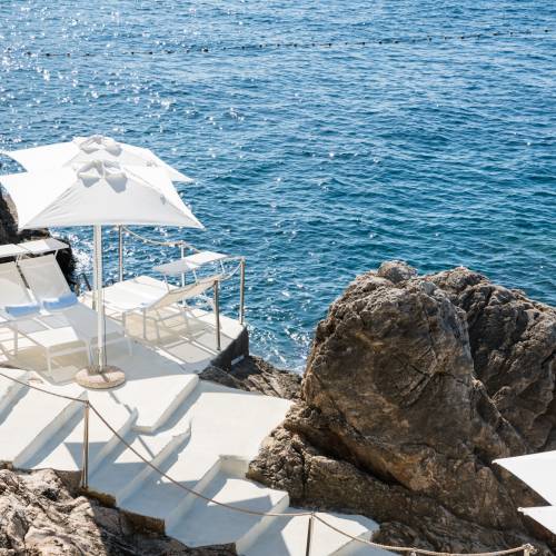 Hotelterrasse mit weißen Liegestühlen und Sonnenschirm direkt am Felsen über dem Meer