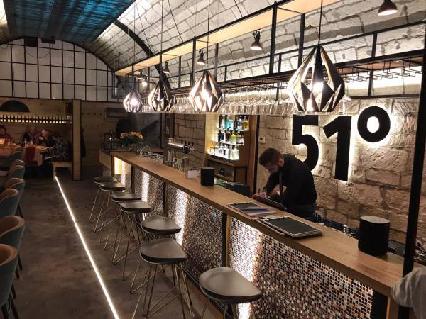 Moderne Weinbar mit einer langen Holztheke, beleuchtet von geometrischen Lampen. Im Hintergrund sind Tische und Stühle sichtbar. Die Zahl '51°' ist prominent an der Wand.