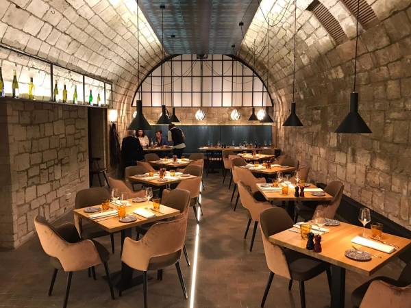 Innenansicht eines Restaurants mit runden Tischen und Stühlen in neutralen Farben. An der Wand sind Flaschen und Lampen sichtbar. Im Hintergrund sind Gäste zu sehen.
