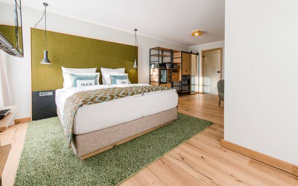 Hotelzimmer mit einem Doppelbett, zwei Kissen mit 'TRAM' Aufdruck, einem Teppich in Grün, Holzfußboden und einer Küchenzeile im Hintergrund.