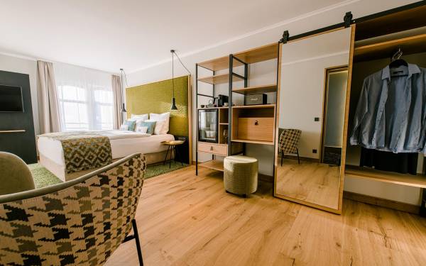 Hotelzimmer mit einem Doppelbett, einem großen Spiegel, einem Kleiderschrank und einem Stuhl. Holzboden und grüne Wanddekoration.