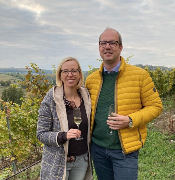 Paar steht in einem Weinberg, die Frau tr&auml;gt eine gestreifte Jacke und h&auml;lt ein Glas Sekt, der Mann tr&auml;gt eine gelbe Jacke und h&auml;lt ebenfalls ein Glas.