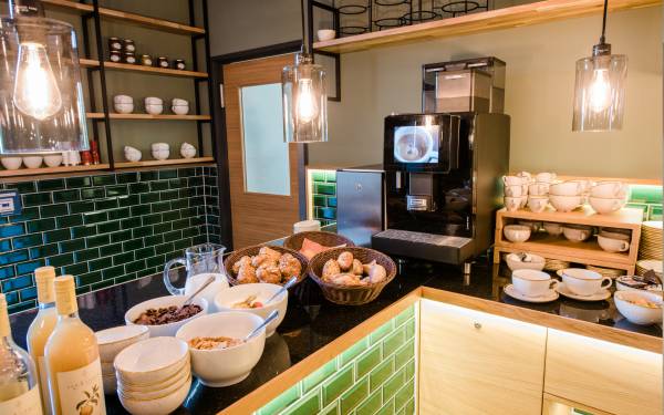 Frühstücksbereich in einer Hotelanlage mit einem Kaffeeautomaten, Schalen mit Croissants und Müsli, Gläsern und Tassen auf einem Tisch.