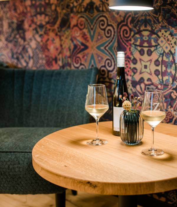 Ein runder Holztisch mit zwei Weingläsern und einer Flasche Wein. Im Hintergrund eine gemusterte Wandtapete und eine kleine Vase mit Blumen.
