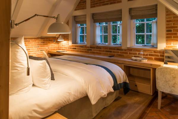 Hotelzimmer mit zwei Betten, weißen Bettdecken und blauen Akzenten. Sichtbare Ziegelwände, Holzfußboden und Fenster mit Vorhängen. Ein Nachttisch mit Lampe und Gläsern.