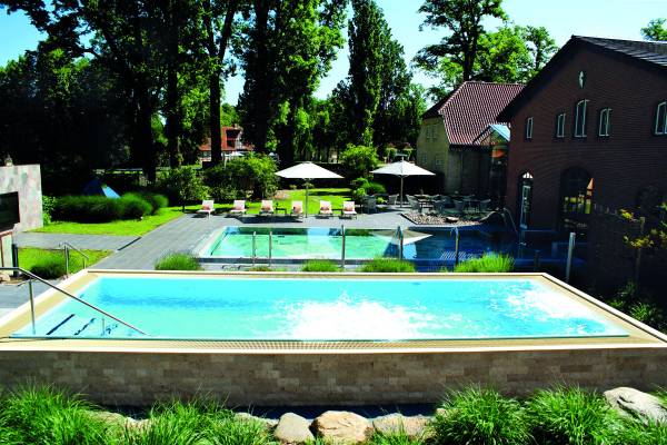 Hotelanlage mit einem Sole-Whirlpool im Vordergrund, umgeben von Rasen und Bäumen. Im Hintergrund sind Liegen und ein weiteres Schwimmbecken sichtbar.