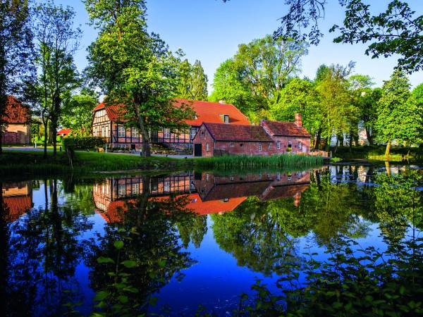 Hotelanlage Weissenhaus mit einem roten Dach und Fachwerk, umgeben von Bäumen und einem Teich, der die Gebäude spiegelt.