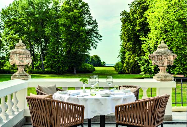 Terrasse eines Hotels mit einem Tisch, vier Stühlen und zwei großen Urnen. Der Tisch ist mit einer weißen Tischdecke und Gläsern gedeckt. Im Hintergrund sind Bäume und eine Wiese sichtbar.