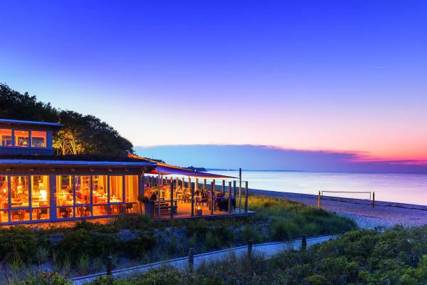 Hotelanlage Weissenhaus mit Bootshaus von Michael Poliza, abendliche Szenerie am Strand, beleuchtete Terrasse, ruhiges Wasser im Hintergrund.
