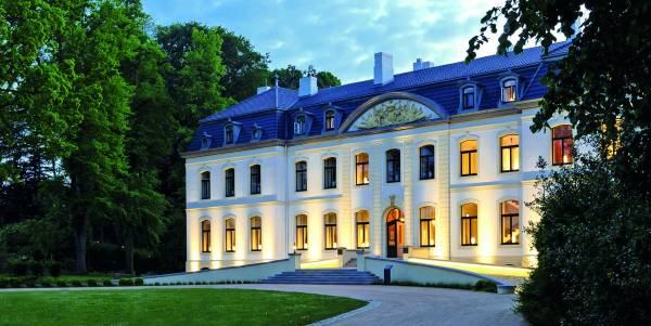 Hotelanlage Weissenhaus mit eleganter Fassade, großen Fenstern und einem geschwungenen Eingang. Umgeben von Bäumen und Rasenflächen.