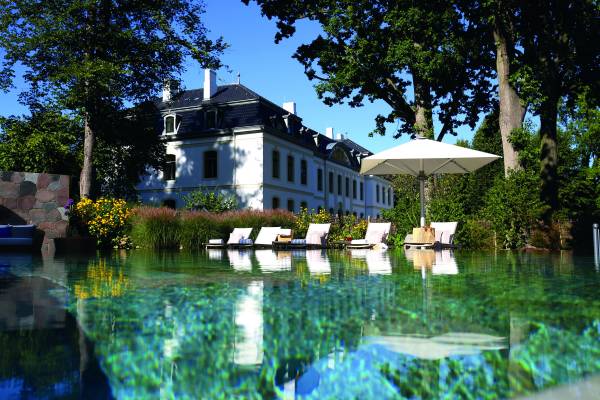 Außenansicht der Weissenhaus Schloss-Therme mit einem Pool und Liegen. Im Hintergrund ist ein historisches Gebäude mit Bäumen und Blumen zu sehen.