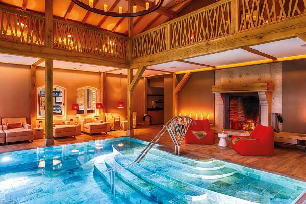 Innenansicht der Weissenhaus Schlosstherme mit einem Pool und Kamin. Der Pool hat eine geschwungene Treppe, umgeben von Holzverkleidung und gemütlichen Sitzbereichen.