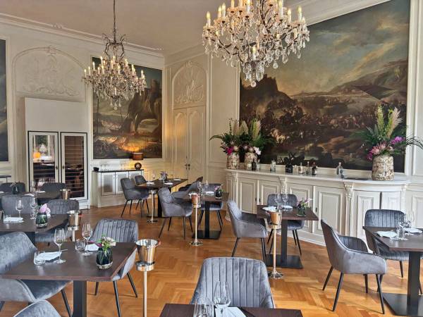 Elegantes Restaurant mit grauen Stühlen, dunklen Tischen, Kronleuchtern und großen Wandgemälden in einem historischen Raum