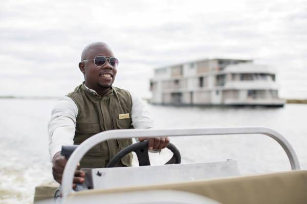 Mann in einem Boot mit Steuerung, trägt eine grüne Weste und ein weißes Hemd. Im Hintergrund ist die Zambezi Queen Hotelanlage sichtbar.