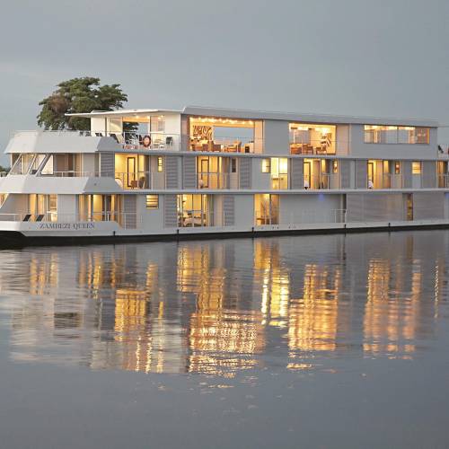 Das Hotelboot 'Zambezi Queen' schwimmt auf ruhigem Wasser, mit mehreren Fenstern, die warmes Licht ausstrahlen. Umgeben von grüner Vegetation.