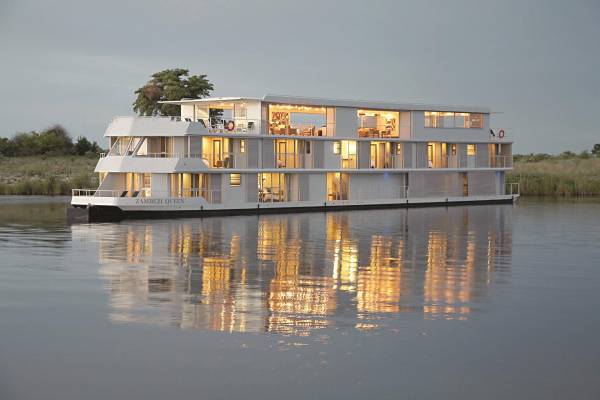 Das Hotelboot 'Zambezi Queen' schwimmt auf ruhigem Wasser, mit mehreren Fenstern, die warmes Licht ausstrahlen. Umgeben von grüner Vegetation.