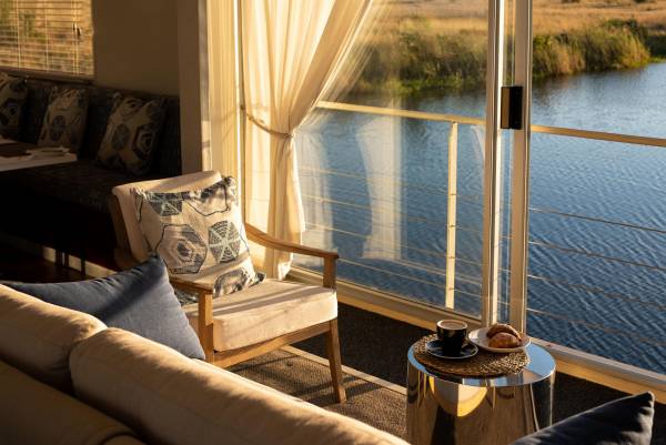 Lounge-Bereich der Zambezi Queen mit einem Sessel, einem Tisch und einem Ausblick auf einen Fluss. Auf dem Tisch stehen eine Tasse Kaffee und ein Gebäck.