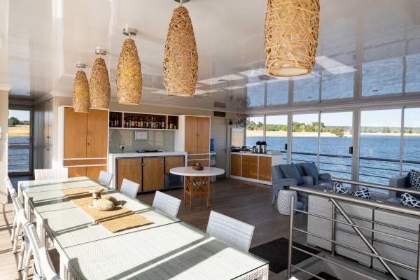 Innenansicht des Speisesaals der Zambezi Queen mit einem langen Esstisch, Stühlen, einer Bar und großen Fenstern mit Blick auf das Wasser.