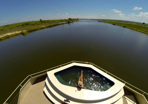 Blick auf den Splash-Pool der Zambezi Queen Hotelanlage mit einer Person, die im Wasser sitzt. Im Hintergrund fließt ein breiter Fluss, umgeben von grüner Landschaft.