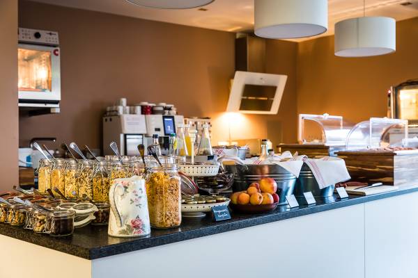 Frühstücksbuffet in einem Hotel mit verschiedenen Gläsern für Müsli, Obst und Marmelade. Ein Kühlschrank und eine Mikrowelle sind im Hintergrund sichtbar.