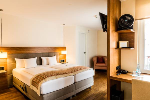 Doppelzimmer in einem Hotel mit zwei Betten, Holzfußboden, einem Sessel, Schreibtisch und Fernseher. Sichtbare Dekoration mit neutralen Farben.
