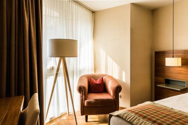 Hotelzimmer mit einem roten Sessel, einem Stehlampe und einem Bett. Helle Vorhänge lassen Sonnenlicht herein.