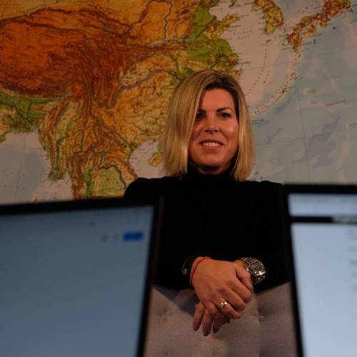 Christine Elsner stehen schwarz gekleidet vor einer großen Landkarte und lächeln freundlich in die Kamera. Im Vordergrund sind links und rechts von ihr Computerbildschirme zu sehen.