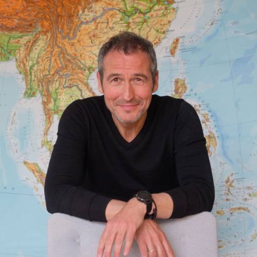 Henrik Heüveldop lehnt mit verschränkten Händen über der Rückenlehne eines hellen Stuhls, er schaut freundlich in die Kamera. Im Hintergrund ist eine großen Landkarte zu sehen.