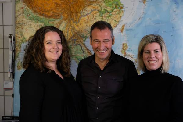 Sharlyn Tripp, Henrik Heüveldop und Christine Elsner stehen schwarz gekleidet vor einer großen Landkarte und lächeln freundlich in die Kamera.